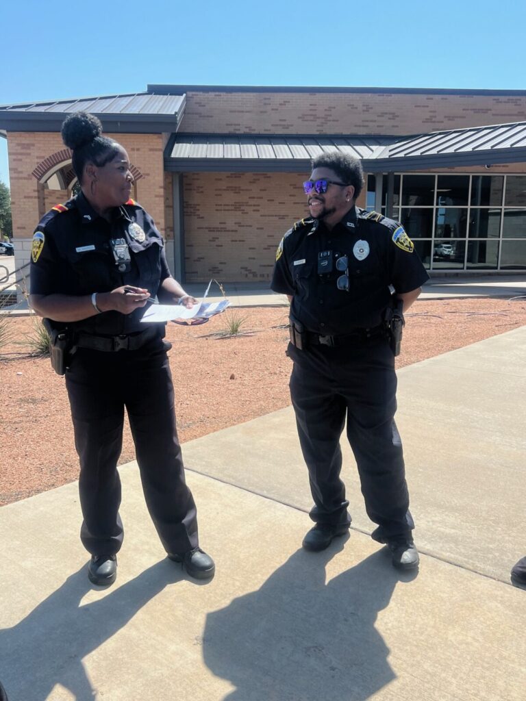 Two police officers in uniform stand outside a building, talking while one holds a clipboard and pen.